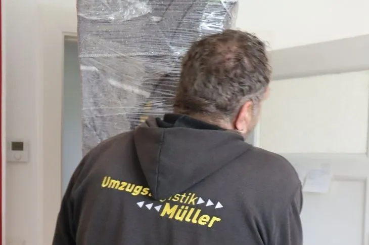umzugshelfer traegt hohes moebelstueck mit schutzfolie